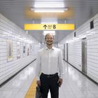 Ein Schild mit "Exit 8" und Aufwärtspfeil in einem U-Bahn-Tunnel. Ein Mann lächelt, trägt Hemd und hält eine Aktentasche.