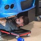 Ein Mechaniker liegt auf einem Rollbrett und arbeitet unter einem blauen Auto in einer Werkstatt. Werkzeug liegt herum.