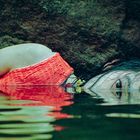 Kein Text zu sehen. Eine Person mit nassem Haar liegt an Felsen im Wasser. Sie trägt ein rotes Oberteil.