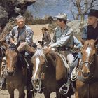 Sieben Männer reiten auf Pferden durch karge Landschaft, tragen Cowboyhüte und Westernkleidung.