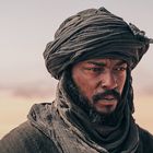 Ein Mann trägt einen dunklen Turban und schaut nachdenklich in die Ferne, umgeben von sandiger Wüstenlandschaft.