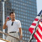 Ein Mann in weißem Poloshirt mit Sonnenbrille am Kragen hält ein Weinglas, daneben weht eine US-Flagge auf einer Yacht.