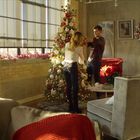 In einem gemütlichen Raum schmücken zwei Personen einen Weihnachtsbaum. Im Raum stehen festliche Dekorationen.