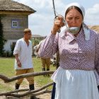 Eine Frau in traditioneller Kleidung trinkt aus einer Metallschöpfkelle, ein Mann in Leinenkleidung hinten.