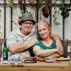 Ein älteres Paar sitzt lächelnd nah beieinander auf einer Bank, umgeben von Pflanzen. Auf dem Tisch sind Bücher und Geschirr.