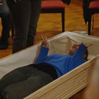 Eine Person in blauer Kleidung liegt in einem offenen Sarg und zeigt mit beiden Händen das Friedenszeichen. Menschen stehen herum.