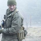 Ein Mann in militärischer Kleidung hält ein Gewehr, steht draußen im Nebel, trägt eine grüne Mütze und Handschuhe.