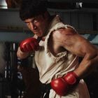 Ein muskulöser Mann in hellem, zerrissenem Outfit mit roten Boxhandschuhen in Kampfpose in einem industriellen Umfeld.