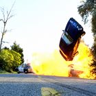 Ein Auto fliegt in der Luft, eine Explosion im Hintergrund, Bäume am Straßenrand, Polizeiwagen auf der Straße.