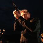 Ein Mann in dunkler Kleidung hält eine Waffe hoch, vor einem dunklen Hintergrund mit schwachem Licht.