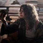 Eine Person mit lockigem Haar hält ein kunstvoll verziertes Objekt in der Hand, umgeben von gestapelten Büchern.