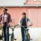 Ein Erwachsener und ein Kind schieben Fahrräder vor einer roten Scheune. Der Mann trägt eine hölzerne Kiste auf dem Fahrrad.