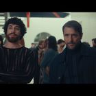 Zwei Männer stehen in einem Raum voller Menschen. Der linke Mann trägt einen durchsichtigen schwarzen Pullover, der rechte einen dunklen Mantel.