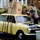 Ein großer Karton mit "TV" ist auf dem Dach eines kleinen gelben Autos befestigt. Ein Mann zieht daran Seile fest.