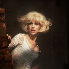 Eine Frau mit blonden, welligen Haaren trägt ein weißes Spitzenkleid und schaut erschrocken hinter einer Ziegelwand hervor.