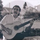 Ein Mann in einem weißen Hemd spielt eine Akustikgitarre im Freien, umgeben von Bäumen und einem unscharfen Hintergrund.