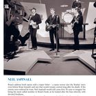 NEIL ASPINALL. Eine Band spielt live auf einer Bühne, umgeben von Kameras. Musiker mit Gitarren und Schlagzeug.