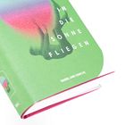 „IN DIE SONNE FLIEGEN“ in weiß auf grünem Hintergrund, mit einem abstrakten Design darunter.