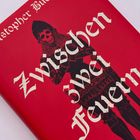 "Zwischen zwei Feuern" steht in großen Buchstaben. Ein Ritter in schwarzem Gewand auf rotem Hintergrund ist abgebildet.