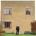 Eine Person steht vor einem zweistöckigen Haus aus gelben Ziegeln mit vier Fenstern, umgeben von Gras und Sträuchern.