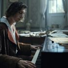 Ein junger Mann in einem eleganten Mantel spielt konzentriert Klavier in einem gemütlichen, lichtdurchfluteten Raum.