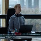 Ein Mann mit Kopfhörern sitzt an einem modernen Glastisch, auf dem ein Laptop und diverse Gegenstände stehen.