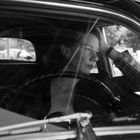 Eine Frau sitzt nachdenklich in einem Auto, schaut aus dem Fenster, Hand am Kopf, Schwarz-Weiß-Fotografie.