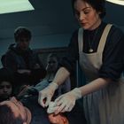 Eine Frau in Schürze versorgt eine Wunde, umgeben von besorgten Menschen in einem schlecht beleuchteten Raum.
