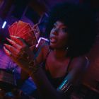 Neon-Schriftzug im Hintergrund einer Bar, Frau mit Afrohaar und auffälligem Schmuck hält Spielkarten in gedämpftem Licht.