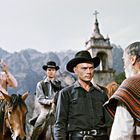 Drei Männer auf Pferden, ein Mann zu Fuß, alle in Cowboykleidung, vor einer Kirche mit Bergkulisse.