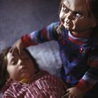 Ein Junge in rot-weißem Hemd liegt mit geschlossenen Augen, während eine Puppe mit grimmigem Gesicht daneben sitzt.