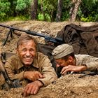 Zwei Männer in historischen Militäruniformen lächeln in einem Schützengraben. Im Hintergrund Bäume und ein Gewehr.