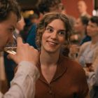 Eine junge Frau mit braunem Haar lächelt in einem belebten Raum. Ein Mann trinkt aus einem Glas.