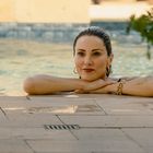 Eine Frau lehnt sich entspannt am Rand eines Pools an. Sie trägt Ohrringe und Armbänder; im Hintergrund sind Pflanzen sichtbar.