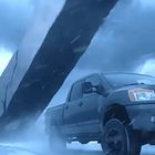 Ein blauer Pickup fährt auf einer Straße. Im Hintergrund wird ein riesiger Lkw-Anhänger in die Luft gewirbelt.