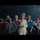 Neun Menschen lachen, während eine Frau in der Mitte ernst bleibt. Der Hintergrund ist dunkel und strukturiert.