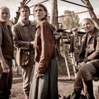 Fünf Personen in historischer Kleidung blicken besorgt in die Ferne. Im Hintergrund sind Bäume und Holzbauten sichtbar.