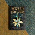 "Wicked Endeavors". Ein Buchcover mit einer Lilie und einem Schwert in der Mitte, blau-goldenes Hintergrundmuster.