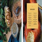 "HOME BAKERY", gefolgt von einer Liste von Backwaren wie "Essential Everyday Dough" und "Homestyle Cinnamon Rolls". Ein Donut mit Glasur, blühende Blumen und Gläser mit Körnern auf Holz.