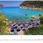 "Anthony Quinn Beach." Sandstrand mit blauen Sonnenschirmen, türkisfarbenes Meer und Felsen, umgeben von grünen Hügeln.