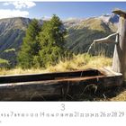 "Alpenbrunnen" steht unten. Holzbrunnen vor alpiner Berglandschaft mit einem Wasserstrahl, umgeben von Tannen.