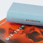 Text: „Sebastian Haffner DIE TOCHTER“. Ein Buch mit hellblauem Umschlag liegt auf einem orangefarbenen Cover.