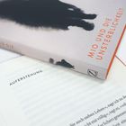 "Buchaufschrift: 'MIO UND DIE UNSTERBLICHKEIT'. Aufgeschlagene Seite: 'AUFERSTEHUNG'. Schwarze, verschwommene Silhouetten."