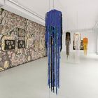 Das Kunstwerk zeigt farbige Stoffskulpturen und eine Wand voll kleiner Gesichter in einem hellen Galerieraum.