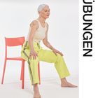 Text: "ÜBUNGEN". Eine ältere Frau mit weißem Haar sitzt auf einem roten Stuhl, trägt gelbe Hose und beiges Oberteil.