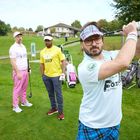 „GOLF COACH“ auf gelbem Shirt. Drei Männer beim Golfspiel auf einem grünen Platz mit Golfbags im Hintergrund.