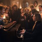 Zahlreiche Menschen in eleganter historischer Kleidung versammeln sich um einen Mann, der Klavier spielt, in einem schwach beleuchteten Raum.