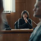 Eine Frau sitzt nachdenklich an einem Tisch in einem Gerichtssaal, andere Personen sind unscharf im Vordergrund.