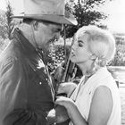 Unten links ist ein Schriftzug. Ein Mann mit Cowboyhut und eine blonde Frau schauen sich nahe an, während sie ein Gewehr hält.