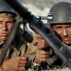 Zwei Soldaten in alten Uniformen, tragen Helme, einer zielt mit einem Gewehr. Sie wirken angespannt und konzentriert.